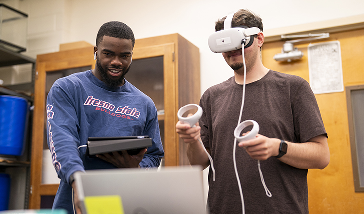 The biology student on the right is wearing a headset and holding a controller while the student on the left is looking at the computer.